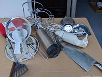 Full view of the mixed kitchen lot on a cardboard surface showing various utensils, hand blender, large cleaver, and other kitchen gadgets.