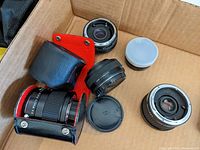 Photograph of four camera teleconverter lenses and a close-up lens, showing black teleconverters and one silver close-up lens, some housed in black and red leather case, displayed inside cardboard box.
