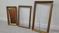 Three antique carved wooden frames displayed against a white wall and light carpet, showing size difference and design.