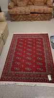 Full view of wool area rug laid on light carpet against background of patterned couch and chair.