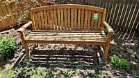Wooden three person outdoor bench shown in a backyard with natural wood color, slatted backrest and seat, flat armrests, weathered condition.