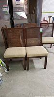 Six wooden dining chairs with cane backs and padded beige fabric seats arranged in two rows in a basement setting. Chairs show visible signs of damage.