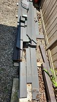 Stack of grey composite decking boards in various lengths stored outdoors on gravel beside a wooden fence