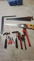 Photo showing the full set of hand tools arranged on the floor including pipe wrenches, hammers, saws and square.