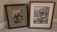 Two framed original art pieces leaning against a wall on carpet. The left piece shows a watercolor painting of houses and trees with sage green matting in a dark wooden frame. The right piece shows a detailed black and white ink drawing with trees and baskets in a medium brown wooden frame.