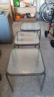 Top and angled view of three square patio side tables with textured glass tops and metal legs lined up on a concrete floor.