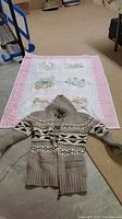 Full view of child's embroidered quilt with pink border and a grey lambswool sweater with geometric patterns laid on floor.