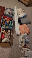 Overview of lot showing bins of thread spools, cones, folded fabric, plastic bags with sewing notions, and vintage sewing patterns.