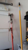 Full view of red extension trimmer and yellow long pole against garage wall.