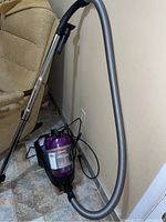Front view of Bissell OptiClean canister vacuum with hose and brush head attached, placed on tiled floor next to couch and wall.