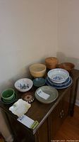 Photo showing all dishes on a wooden cabinet against a wall. Several blue and white porcelain dishes, wooden bowls, a wicker covered basket, and clay serving bowls.