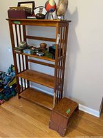 Front view of wooden open bookshelf with three shelves decorated with various small items; small wooden foot stool positioned next to bookshelf.
