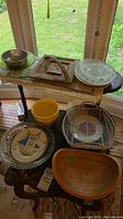 Photo showing multiple bowls and trays arranged on tables including wood bowls, glass bowls, a metal mesh tray, and a clay tray.