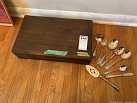 Closed wooden case holding the silverplate cutlery set, with some serving utensils placed beside it on the floor.