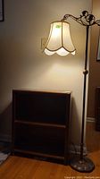 Full view of floor lamp with fabric shade next to 2-tier wood bookcase against wall.