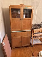Full view of the wooden cabinet with two glass doors on top and two wood doors below with one drawer in between, standing on a hardwood floor.