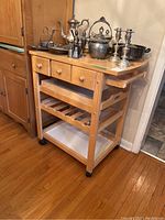 Full view of wood kitchen cart with silver items on top, showing three drawers, towel rack on side with damage, and caster wheels.