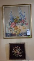 Framed needlepoint floral artworks hanging on wall, larger piece with glass above smaller piece without glass.