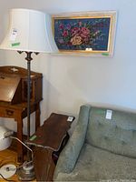 Wide angle shot showing framed floral needlepoint artwork above the vintage wooden side table with magazine rack and nearby floor lamp and furniture.