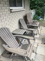 Two wooden Muskoka chairs with a natural, weathered finish and two accompanying small wooden side tables placed on a stone patio against a brick wall.