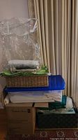Stack of different storage items including a clear plastic bag, green fabric linen on top of a wicker basket, a large blue plastic container, and boxes stacked together, holding assorted table linens.