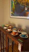 Photo shows all 9 teacups with matching saucers displayed on a wooden cabinet, showcasing a range of color and decorative styles.
