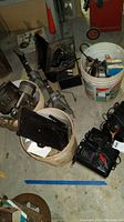 Photo showing buckets with various metal machine parts and power drill, metallic jack stand nearby, items cluttered on basement floor.