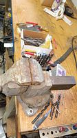 Rusty vintage bench vise mounted on wooden workbench with assorted drill bits and hand tools arranged around it.