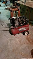 Photo showing entire red Sears air compressor with two cylinders, black motor, wheels, and handle on a concrete basement floor.