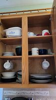 Kitchen cabinet with shelves holding white ceramic baking dish with blue floral pattern and multiple plates, bowls, and mugs in various colors and sizes.