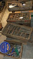 Overview of several wooden trays filled with assorted copper plumbing fittings, nuts, and bolts, plus floor dollies in basement setting.