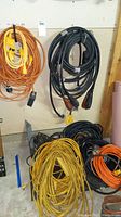 Multiple coiled extension cords in orange, black, and yellow colors hanging on wall and laying on floor.