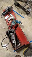 Red toolbox with multiple power tools including air grinder, screwdriver, reciprocating saw, and another power tool amidst cords and wires on concrete floor.