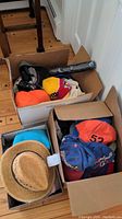 Three cardboard boxes on wood floor each filled with assorted hats and caps, different styles visible