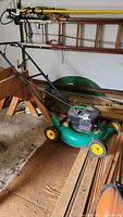 Full view of Weed Eater lawn mower showing green body, yellow wheels, handle, and stored in garage with wooden planks nearby.
