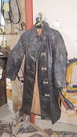 Full view of the vintage fireman leather jacket hanging, showing black leather exterior with metal clasp closures and tan interior lining.
