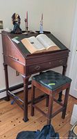 Wooden Palmer Home Collection drop-front desk with open book on top and decorative figurines behind it. Front drawer with metal handles and four legs visible.