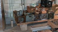 Wide shot showing multiple crates filled with rusted plumbing parts, metal pipes, hooks, chains, and boxes in a basement corner.