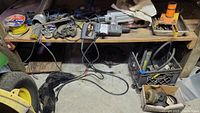 Overview of various power tools and accessories laid out on wooden shelves and floor including drills, saws, welding rods, wiring, and parts in crates and boxes.