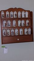 Front view of wooden spice rack mounted on wall holding 21 ceramic mortar-shaped spice containers with Hummel artwork and labeled spices.
