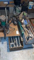 Overview photo showing hydraulic power pump with coiled hoses, disc grinder, wooden tool box filled with hand tools, all on a wooden surface in basement area.