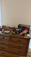 Wide shot of assorted toy cars displayed on wooden dresser, varying in style, size, and material.