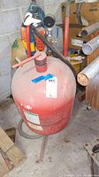 Front angle showing red cylindrical sand blaster tank with pressure gauge and control valves, standing on three legs, with seller lot tag attached.