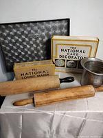 Vintage baking lot arranged with two wooden rolling pins, two labeled boxes for cookie maker and cake decorator, and a metal baking pan.