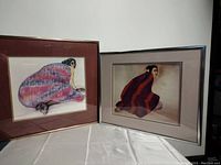 Two framed art pieces side by side, showing women seated and wrapped in colourful striped blankets, each framed with mats and simple frames.
