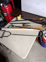 Photo of assorted garage and yard tools on a table showing manual post hole digger, hand saw, crowbar, shovel handle, and more.
