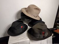 All four vintage men's felt fedora hats shown together on a white surface against a wall, showcasing different colors and bands with feather accents.
