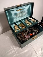 Open green Mele jewelry box showing satin lining and assortment of costume jewelry including brooches and necklaces.