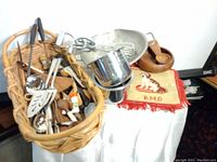 Overall view of the basket containing various metal kitchen utensils including ladles, spatula, tongs, and a whisk, alongside a metal colander and wooden items.