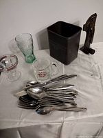 Wide view of the flatware set, glassware, metal container, and bird-shaped bottle opener on white cloth